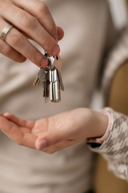 A close-up of a person holding a silver keychain shaped like a house against a blue background, with another person with glasses and facial hair observing in the background. The scene appears to be in